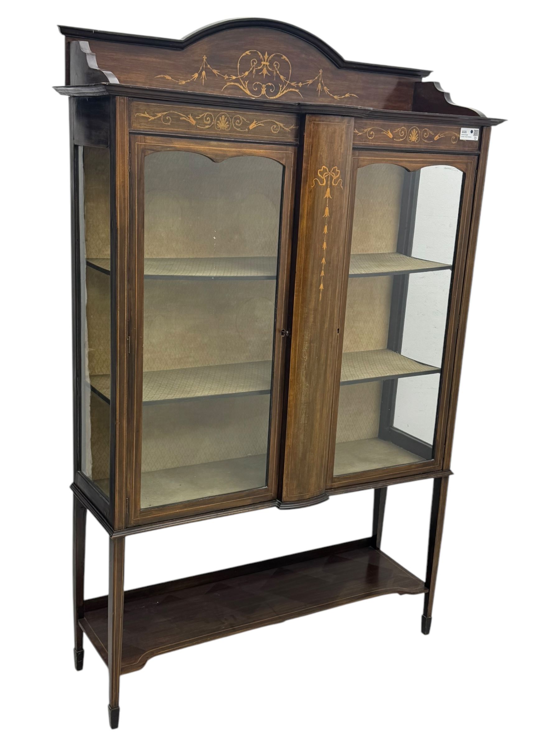 Edwardian mahogany and satinwood crossbanded display cabinet, arched pediment centred by marquetry scrollwork and ribbon decoration, twin glazed doors enclosing a lined a shelved interior, raised on square tapering supports joined by a shaped undertier with diamond parquetry inlay, terminating in spade feet
