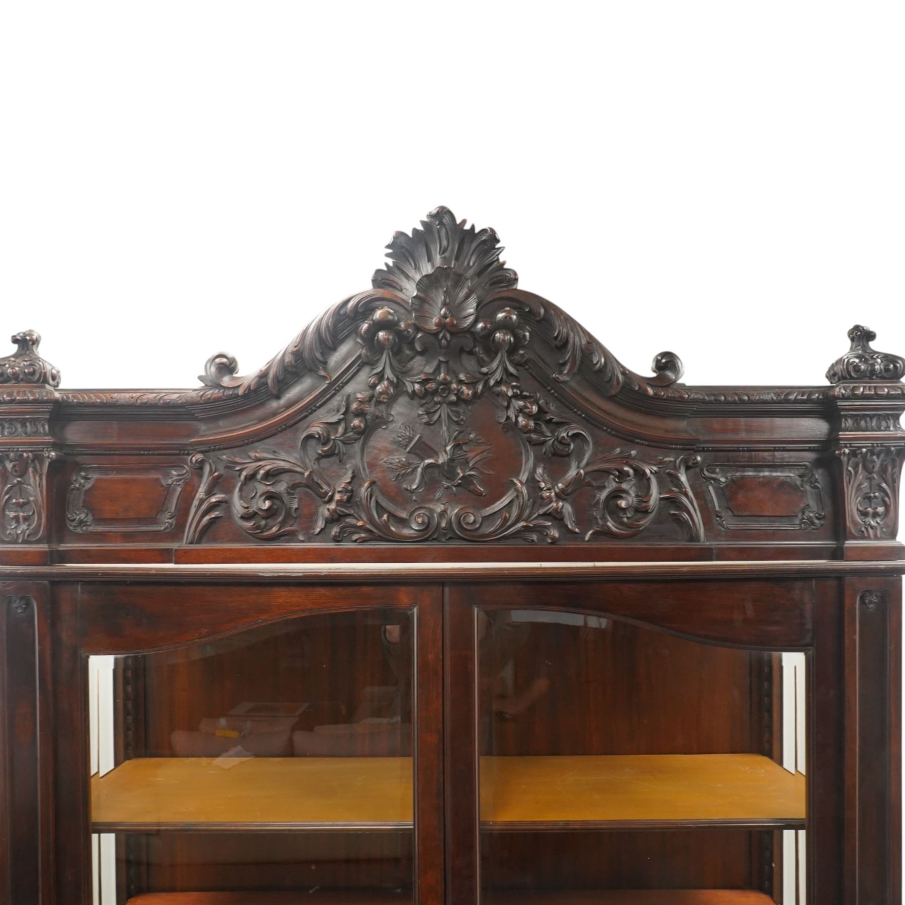 Large and impressive 19th century heavily carved mahogany display cabinet, the ornate pediment carved with a central scalloped and foliate rocaille motif with extending acanthus leaves and egg-and-dart moulding flanked by acanthi finials, over a central cartouche with carved bow-and-arrow motif and trailing scrollwork, the bookcase enclosed by two glazed doors with bevelled glass sides, the base with a gadrooned upper edge over a central rocaille ornament with scrolling acanthus leaves, the panelled frieze with further foliate motifs, raised on large carved snarling lion mask supports with floral garlands and claw feet, united by a concave rectangular platform base with gadrooning egg-and-dart moulding
