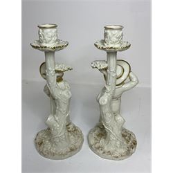 Pair of Royal Worcester figural candlesticks designed by James Hadley in the style of Kate Greenaway, modelled with a young boy and girl with painted features beside stylised ivy clad tree stumps supporting naturalistic sconces and drip pans, raised upon rocky circular bases, decorated with gilt detail throughout, with printed puce stamped marks and impressed 1141 beneath, H26.5cm