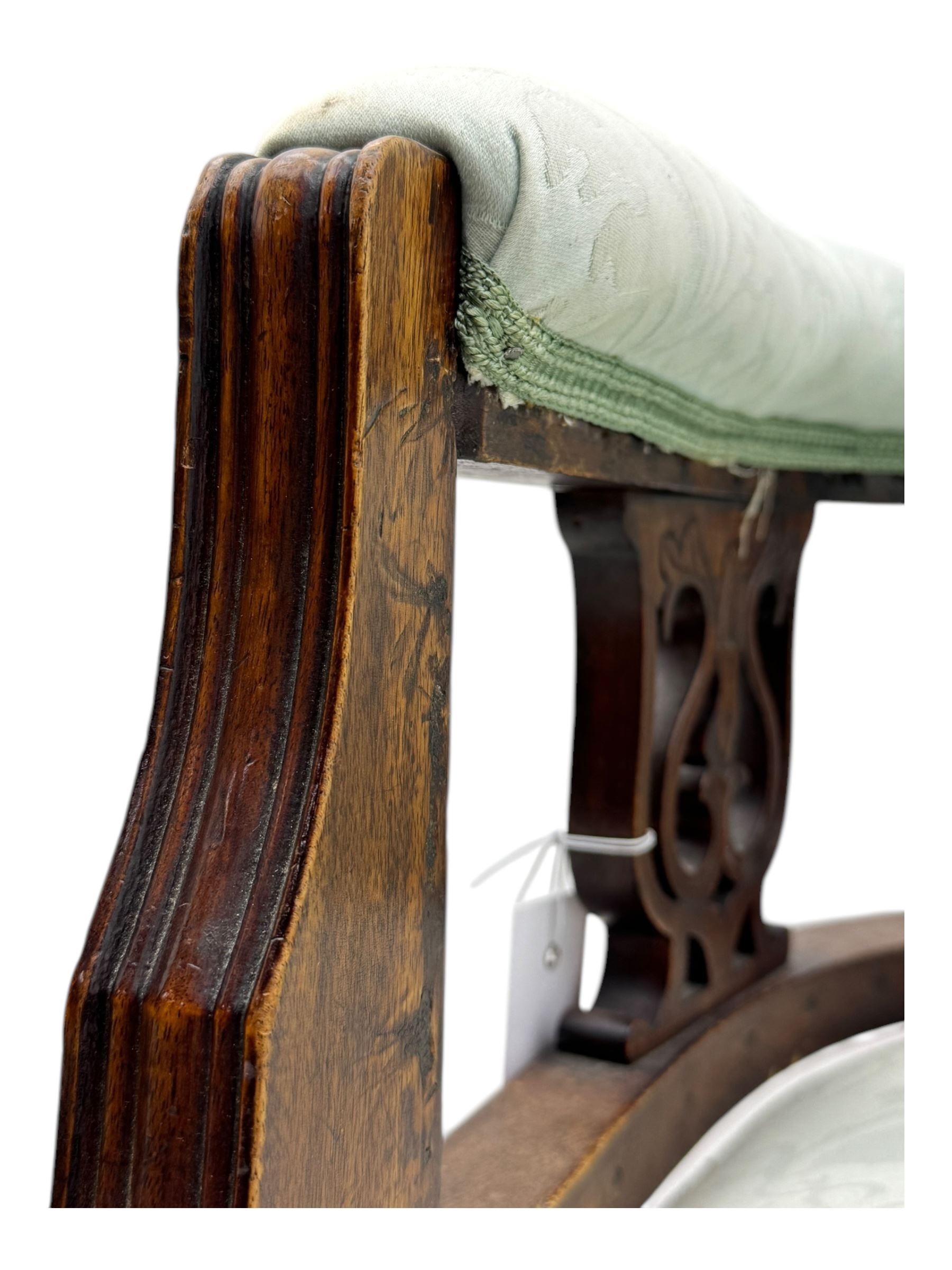 Late Victorian carved mahogany open armchair, the shaped back with fluted upholstered panel and padded arms above pierced scroll carved side supports, over a broad upholstered seat, on turned front supports