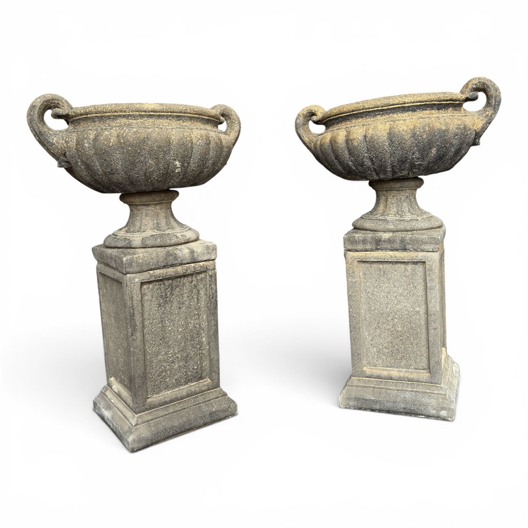 Pair of Neoclassical design weathered cast stone garden urns of kylix form, the rolled rim flanked by twin pierced scroll handles over a shallow gadrooned bowl, raised on a fluted circular socle and faux panelled square pedestal base