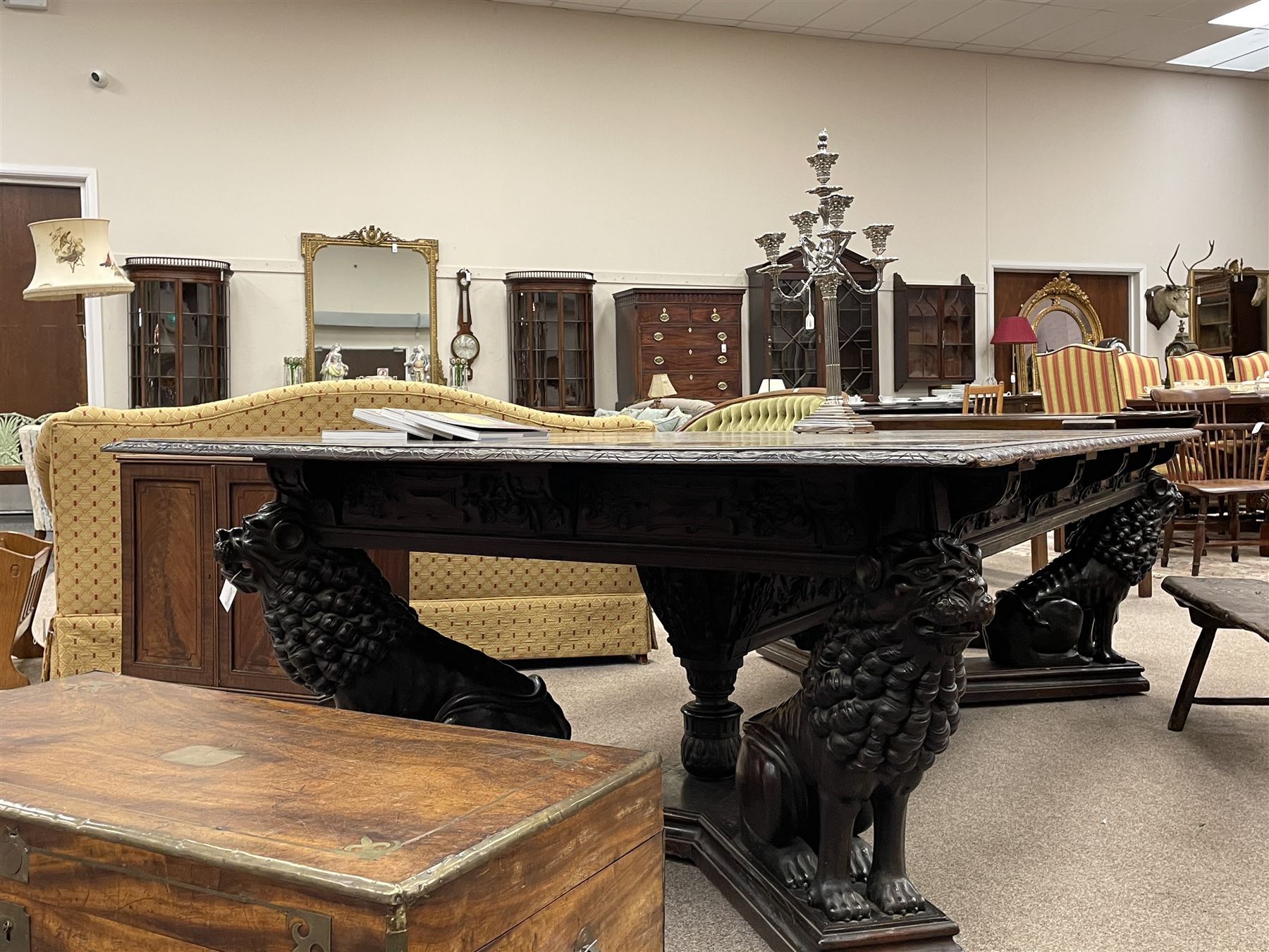 Large 19th century carved oak library table, the rectangular top with lunette carved edge over cartouche panelled and carved  scroll frieze, the central tapering rectangular support relief carved with masks and garlands, on four carved seated lion supports at each corner, on flat angular stretcher with recessed castors stamped ‘Bartholomew & Fletcher, London’