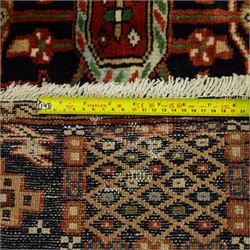 North West Persian Karajeh hand-knotted rug, terracotta ground, with a stepped geometric pole medallion design in blue, ivory and dark navy tones, surrounded by angular tribal motifs incorporating stylised flowerheads, hooked devices and geometric vine work, enclosed by a dark navy border with repeating rosettes and angular leaf motifs, flanked by narrow patterned guard stripes, with cotton fringes