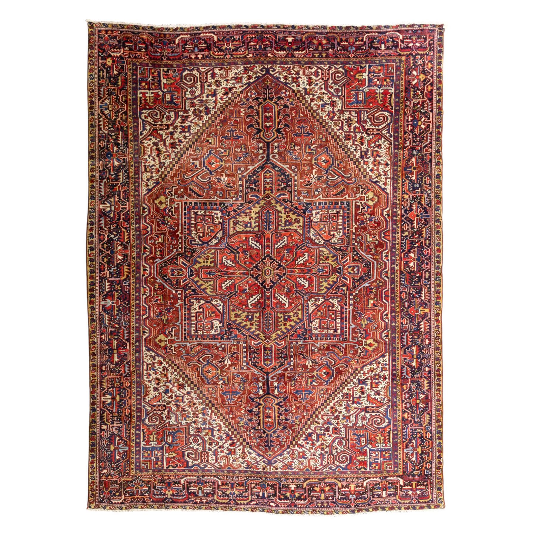 North West Persian Heriz hand-knotted carpet, madder red field, with a large angular stepped central medallion with serrated outlines, flanked by bold stylized palmettes, hooked leaves and geometric devices, spandrels similarly decorated within a wide geometric main border featuring angular floral motifs, enclosed by multiple guard borders in madder red, indigo blue, ivory, olive green and soft ochre, with cotton fringe ends 