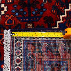 Central Persian Joshagen hand-knotted carpet, madder-red field with a central stepped lozenge medallion and densely filled with stylized geometric floral and tree motifs arranged in a repeating lattice enclosed by indigo spandrels and a broad navy border of angular palmette and vine ornament, flanked by multiple guard stripes, cotton fringe ends 