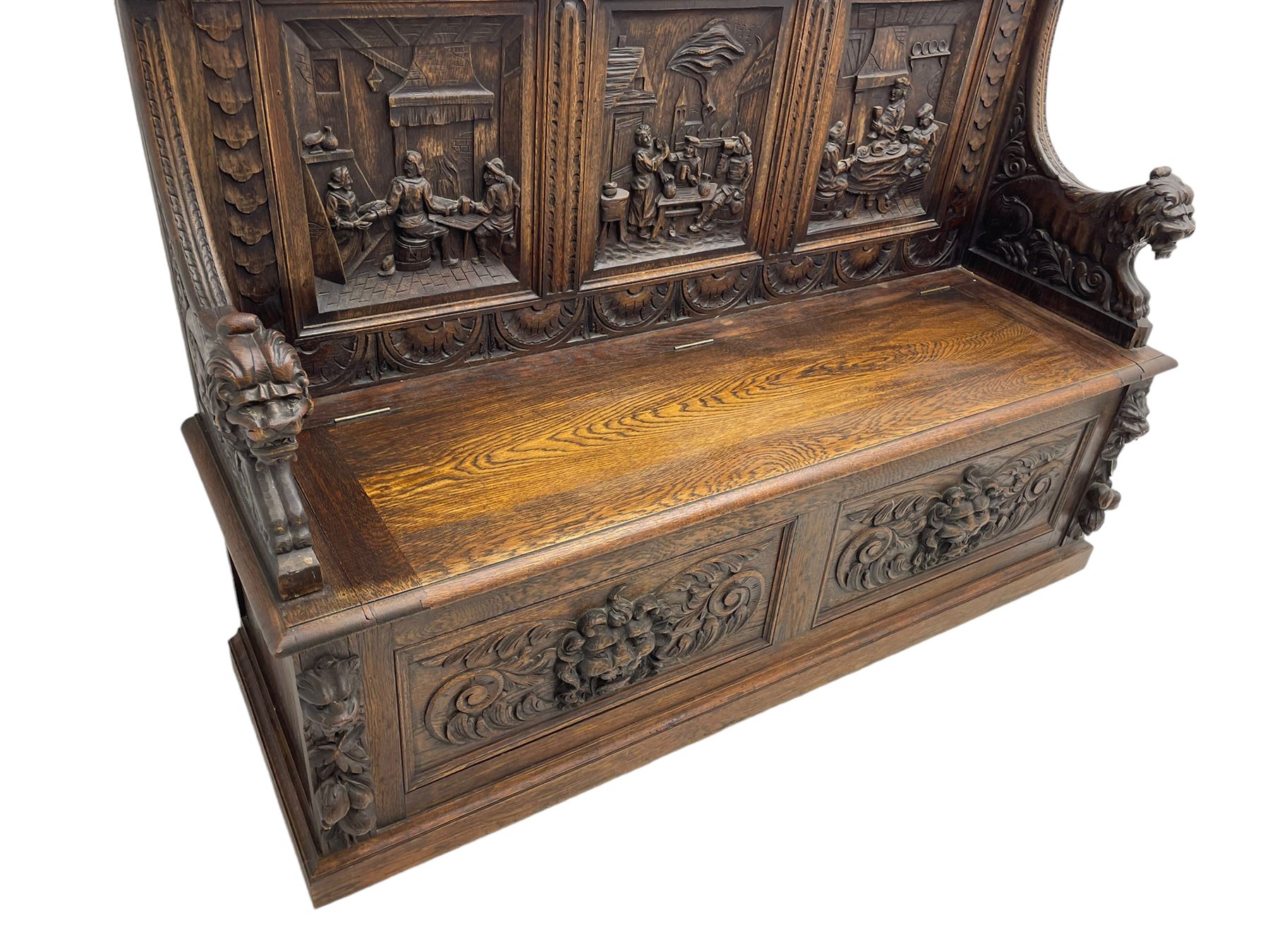 19th century heavily carved oak settle, the moulded upper rail over triangular foliage carved frieze, three Flemish style carved panels depicting tavern scenes, acanthus carved uprights terminating to lion carved arms, hinged box seat, the panelled front carved with lion masks and extending scrolled acanthus leaves, on moulded skirt base