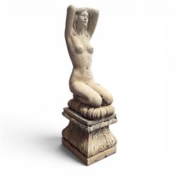 20th century composite stone garden figure, modelled as a kneeling nude female with arms raised behind her head, mounted on an integral cushion, the squared ogee pedestal moulded with acanthus leaves and flower heads, in a white painted and weathered finish