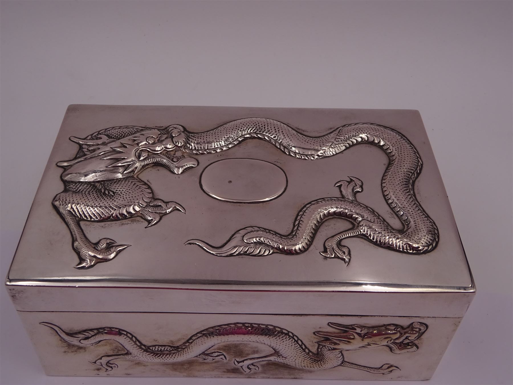 Late 19th century/early 20th century Chinese export silver mounted cigarette box, of rectangular form, decorated in relief with dragons, the hinged cover with similar dragon decoration and central blank oval cartouche, opening to reveal a softwood lined compartmentalised interior, stamped beneath with character mark and KMS for maker Kwong Man Shing, H6.3cm, W15.5cm, D9.5cm