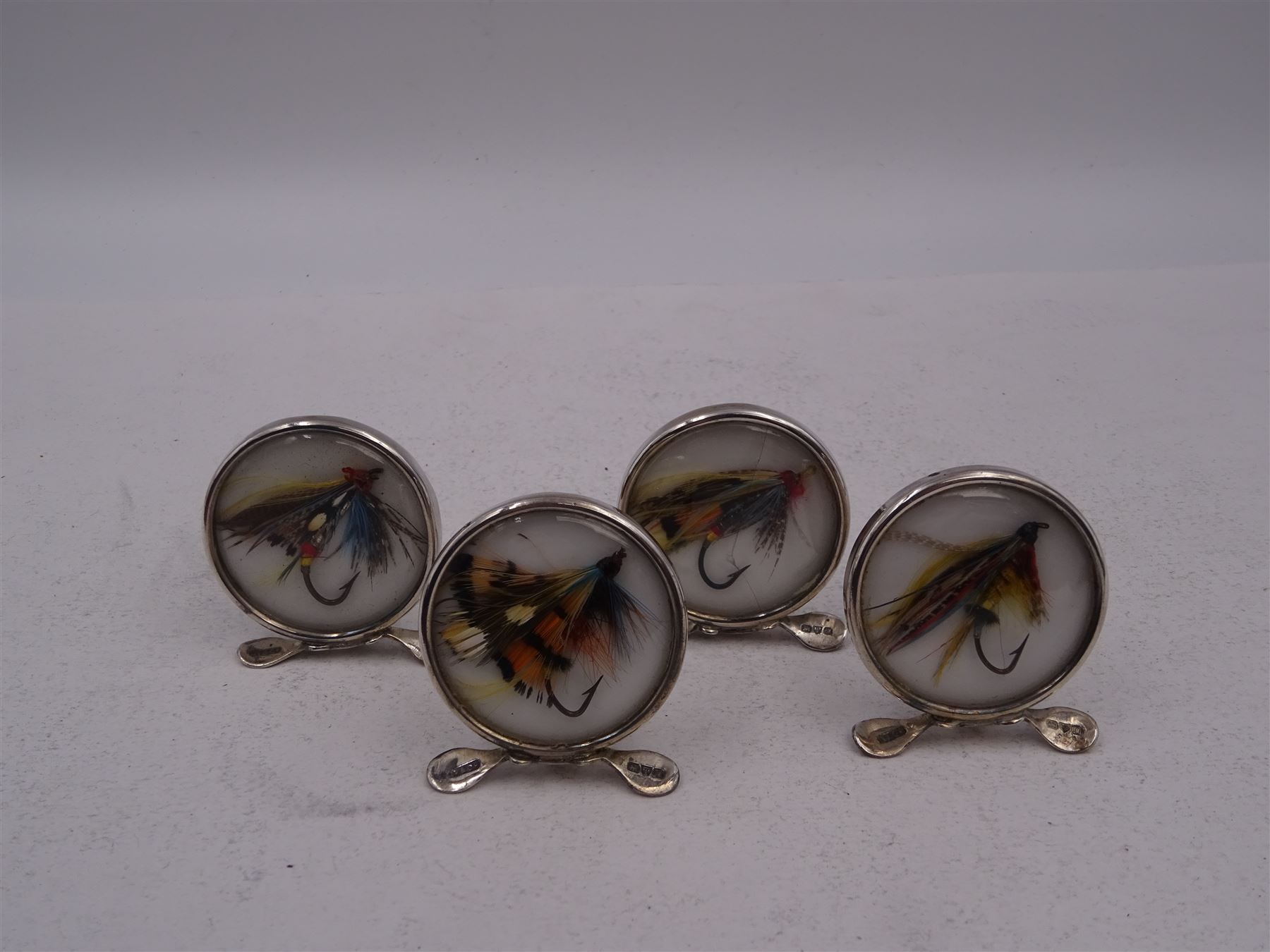 Set of four early 20th century silver mounted place card holders, each of circular form and set with a coloured fly fishing hook behind a glazed panel, upon three pad feet, hallmarked Grey & Co, Chester 1912, H4.5cm, contained within red fitted case, with purple silk and velvet interior