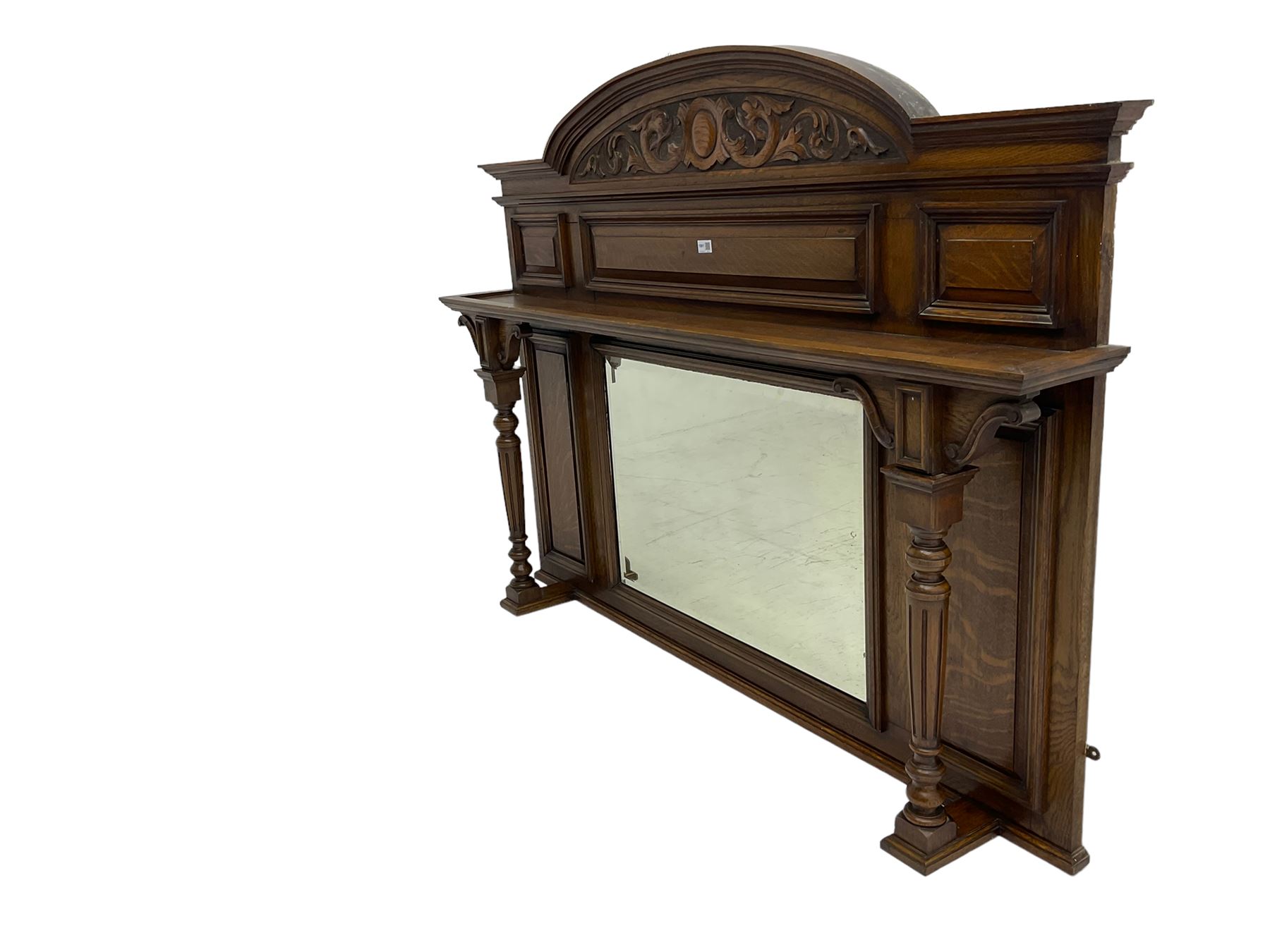 Oak overmantle mirror, arched pediment carved with shield and flanking fish with trailing scrollwork, panelled frieze over extending canopy raised by turned and fluted column pilasters, bevelled mirror plate 