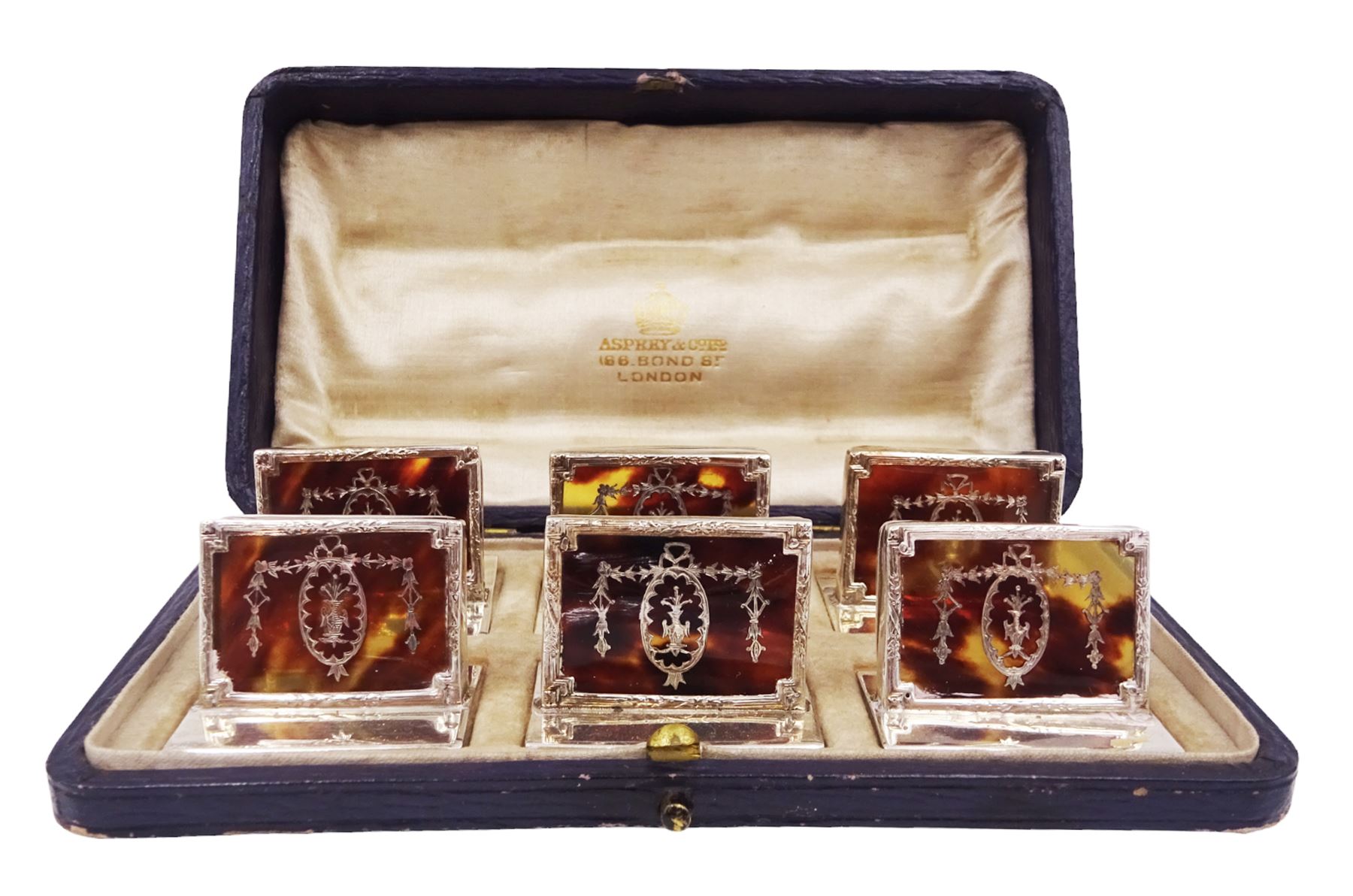 Set of six early 20th century silver mounted tortoiseshell place card holders by Asprey, the rectangular silver reed and ribbon frame bordering a central tortoiseshell panel inlaid with a silver floral motif, upon an oblong foot, hallmarked Asprey & Co Ltd, London 1913, H3cm, contained within a tooled leather fitted case with velvet and silk lined interior 
