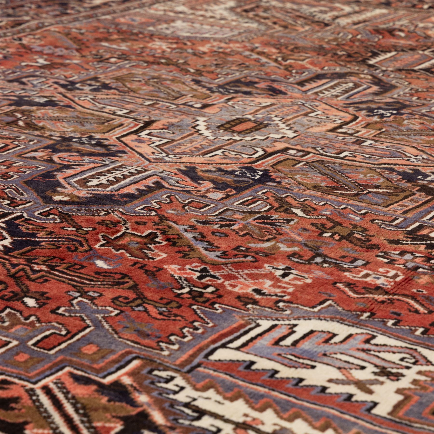 North West Persian Heriz hand-knotted carpet, madder red field with central stepped geometric medallion with radiating angular motifs and serrated outlines, and stylized floral and tribal devices framed by a broad border of repeating angular palmettes and rosettes flanked by multiple guard stripes, with cotton fringe ends 