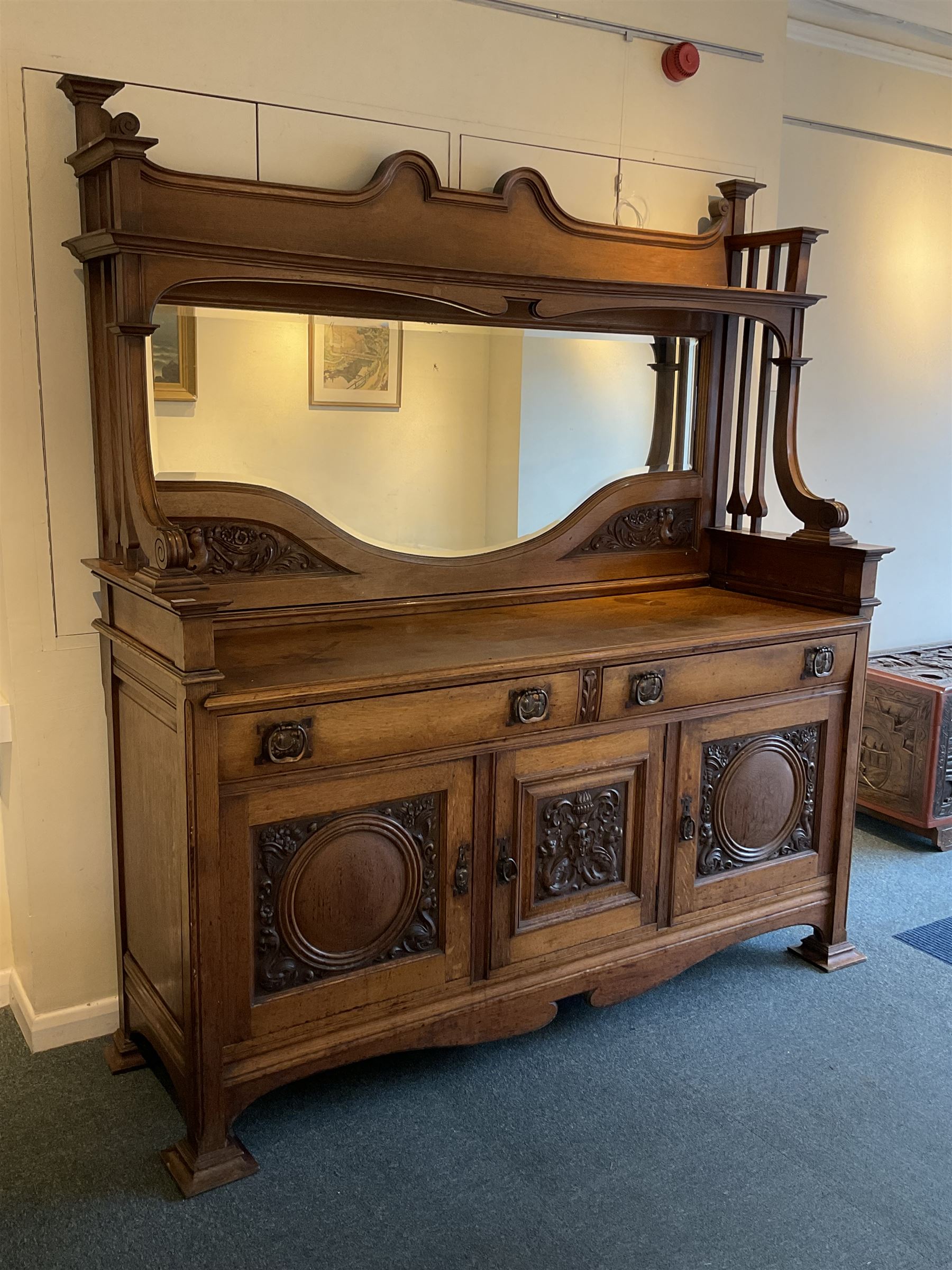 Late 19th century Arts & Crafts period oak mirror back sideboard, shaped bevelled mirror back carved with foliate scrolls and flower heads, down swept supports with elongated club-shaped uprights, moulded rectangular top over two drawers and three cupboards, the drawer fronts with copper handles decorated with sailing ships, panelled doors carved with mythical beasts, flowerheads and foliage, on moulded block feet