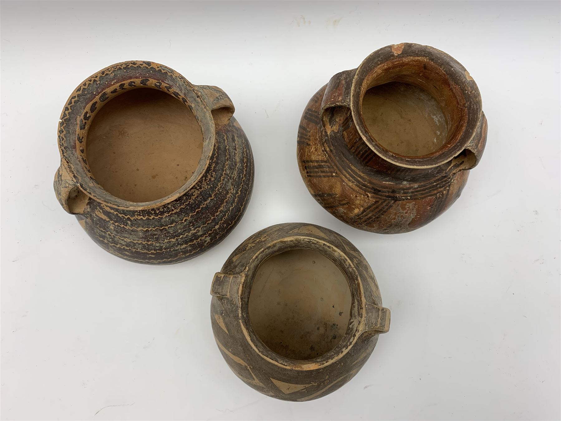Three Chinese Neolithic pottery vessels, each with twin loop handles to the shoulder and painted geometric banded decoration to the body, all bearing labels for Eskenazi London, tallest H14cm