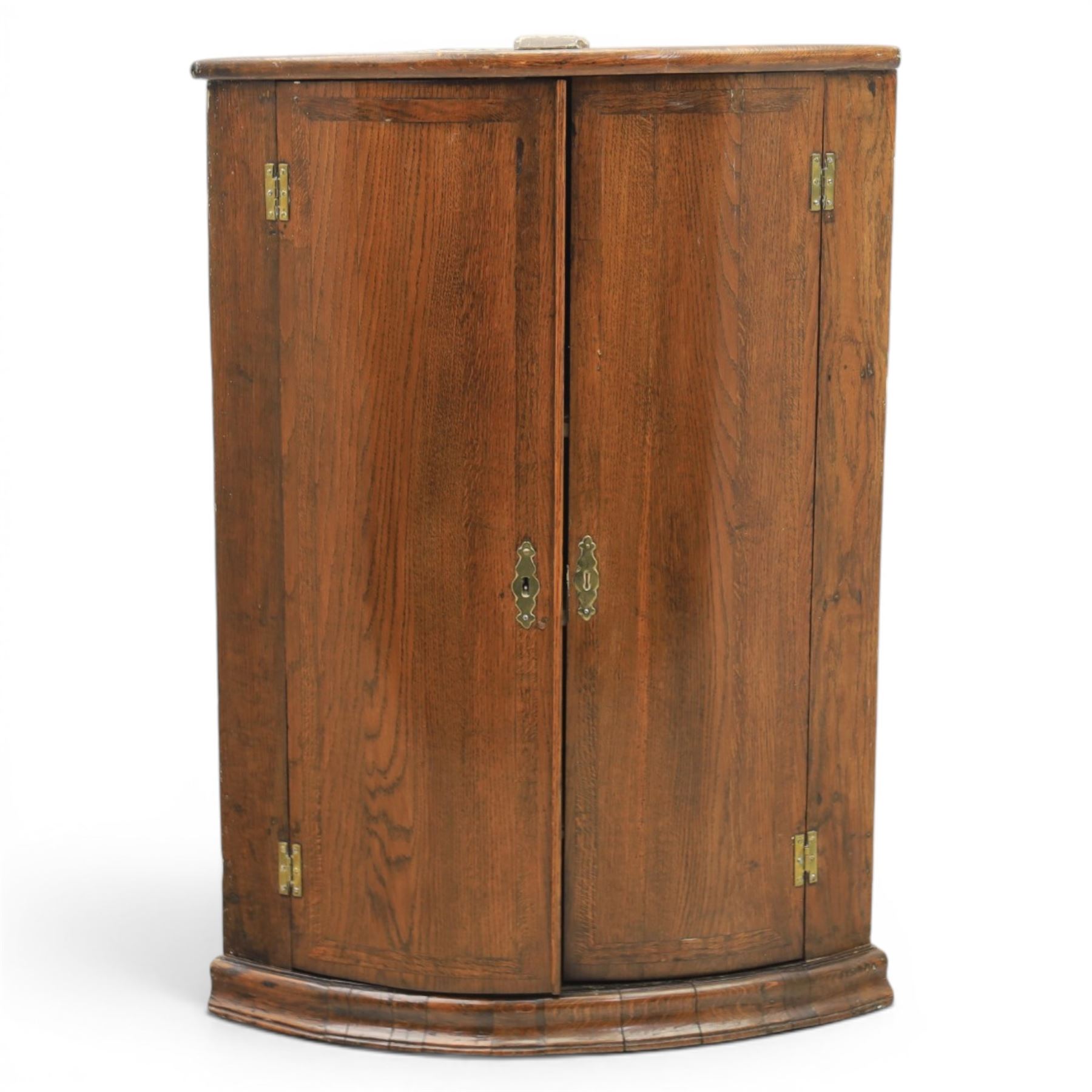 George III oak cylindrical corner cupboard, bow fronted doors revealing a painted shelved interior, brass escutcheons  and hinges, raised on a moulded plinth base