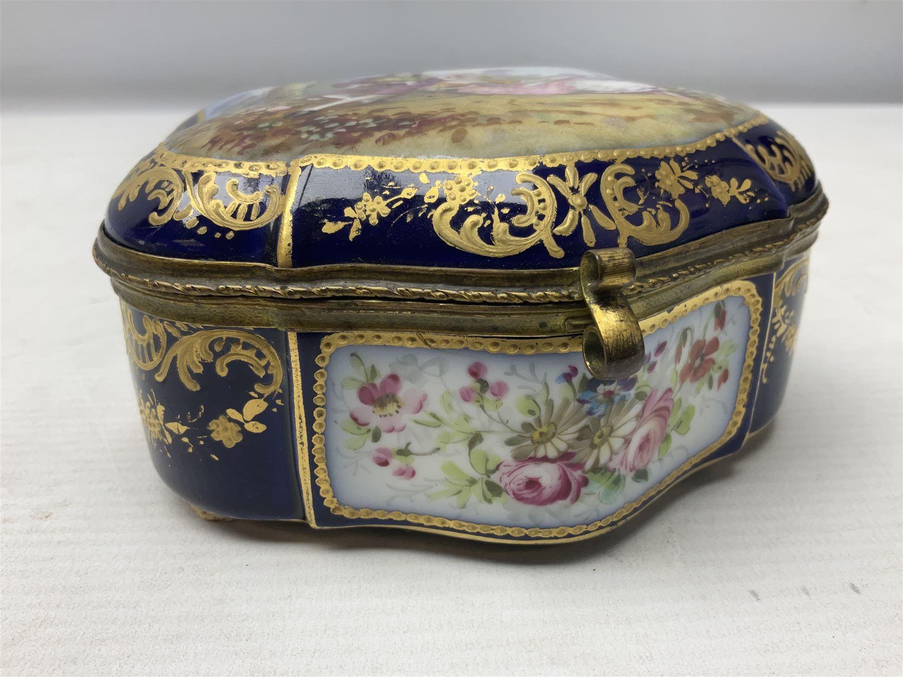 Sevres style late 19th/early 20th century trinket box decorated in the rococo style, the shaped cover painted with an 18th century courting couple in a garden scene opening to reveal the interior painted with floral sprays, the conforming body with a painted panel of floral decoration, all reserved upon cobalt blue ground with foliate gilt borders, with interlaced L's mark beneath, L12cm H6cm