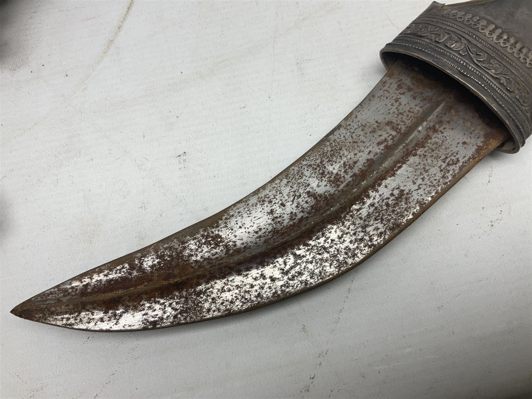 Omani Khanjar dagger, the 19cm curved blade with pronounced medial rib to either side, waisted hilt with dome shaped pommel and silver mounted horn grip decorated with an embossed band; in an intricately decorated silver scabbard with silver rings woven into the belt mount and fitted with original coin mounted silver thread-work belt with additional side knife in sheath verso L34cm overall, belt length 81cm; together with a Nepalese kukri in leather scabbard with one skinning knife (2)