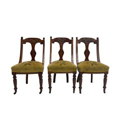 Set three early 20th century mahogany dining chairs, splat back carved with scrolling foliate design, sprung seat upholstered in olive green floral patterned fabric, raised on turned supports with ceramic castors 