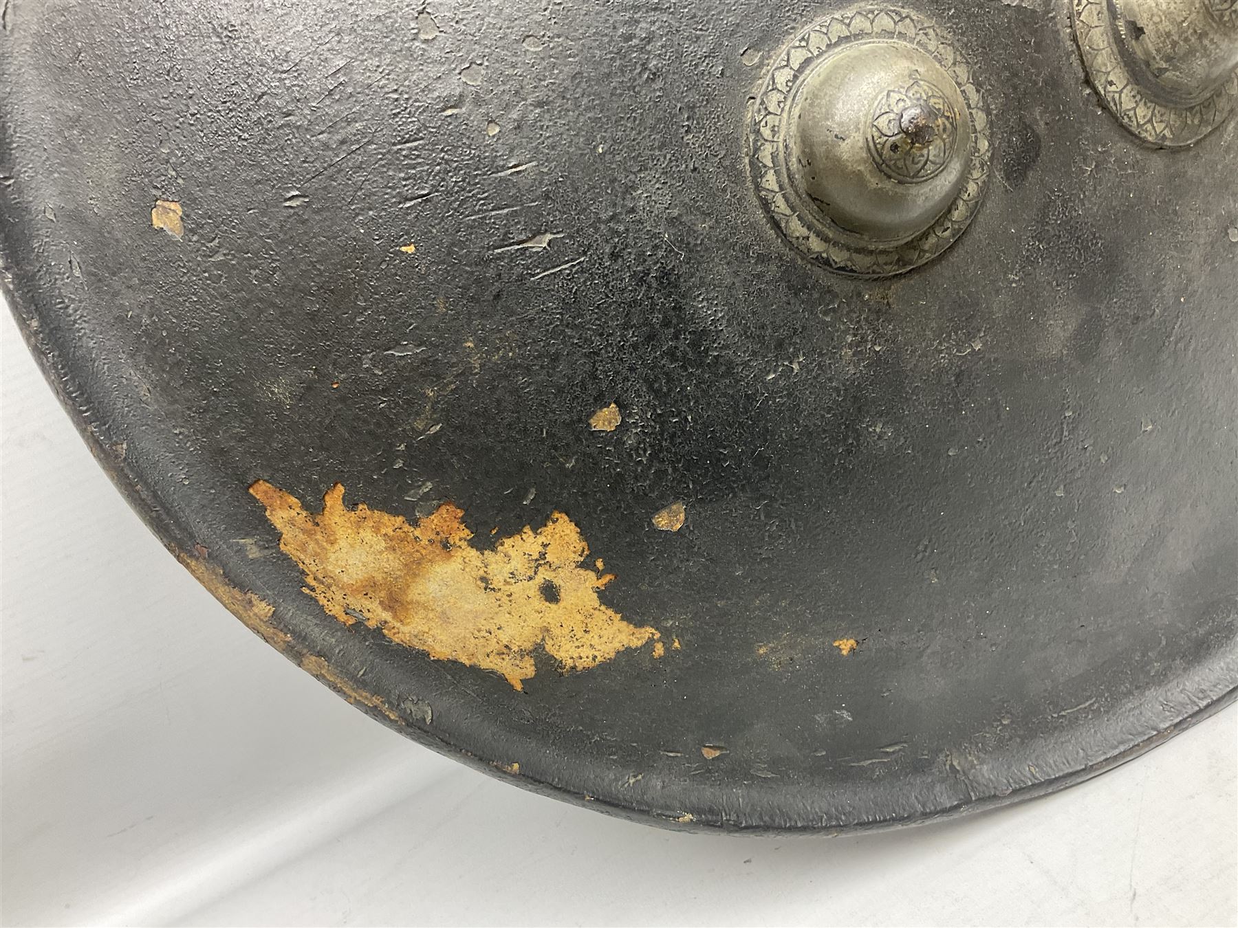 Indian black lacquered hide shield/dhal, of circular convex form with strongly outward-turned brim, fitted at its centre with four metal bosses, each of hemispherical form, with traces of silvered foliate decoration, the bosses corresponding on the inside with four iron rings for enarmes D49cm