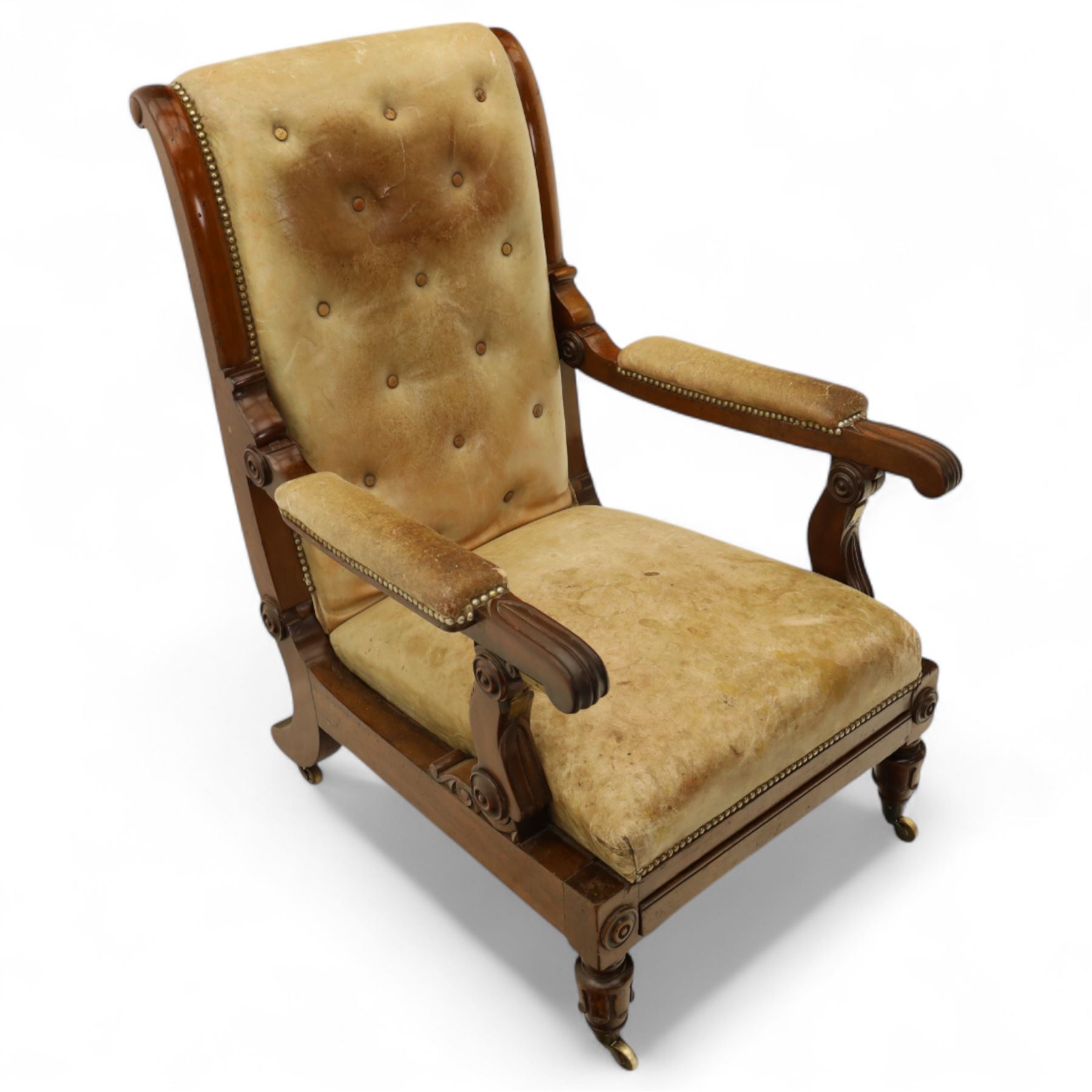 William IV mahogany gentleman's library reclining armchair, leather button upholstered and brass-studded back and seat, above leather-covered arms terminating with carved roundels and foliate scroll-carved ends, underside with brass mechanical trigger reclining lever, the apron with retractable foot rest, supported on lappet carved and turned legs with brass castors, circa 1835