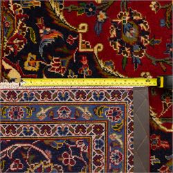 Central Persian Kashan hand-knotted carpet, crimson ground, with a central shaped palmette medallion with pendants, surrounded by scrolling foliate and floral motifs incorporating rosettes, palmettes, and vine work, enclosed by a dark navy with repeating border with repeating palmettes and meandering vines, flanked by floral guard stripes, with cotton fringes 