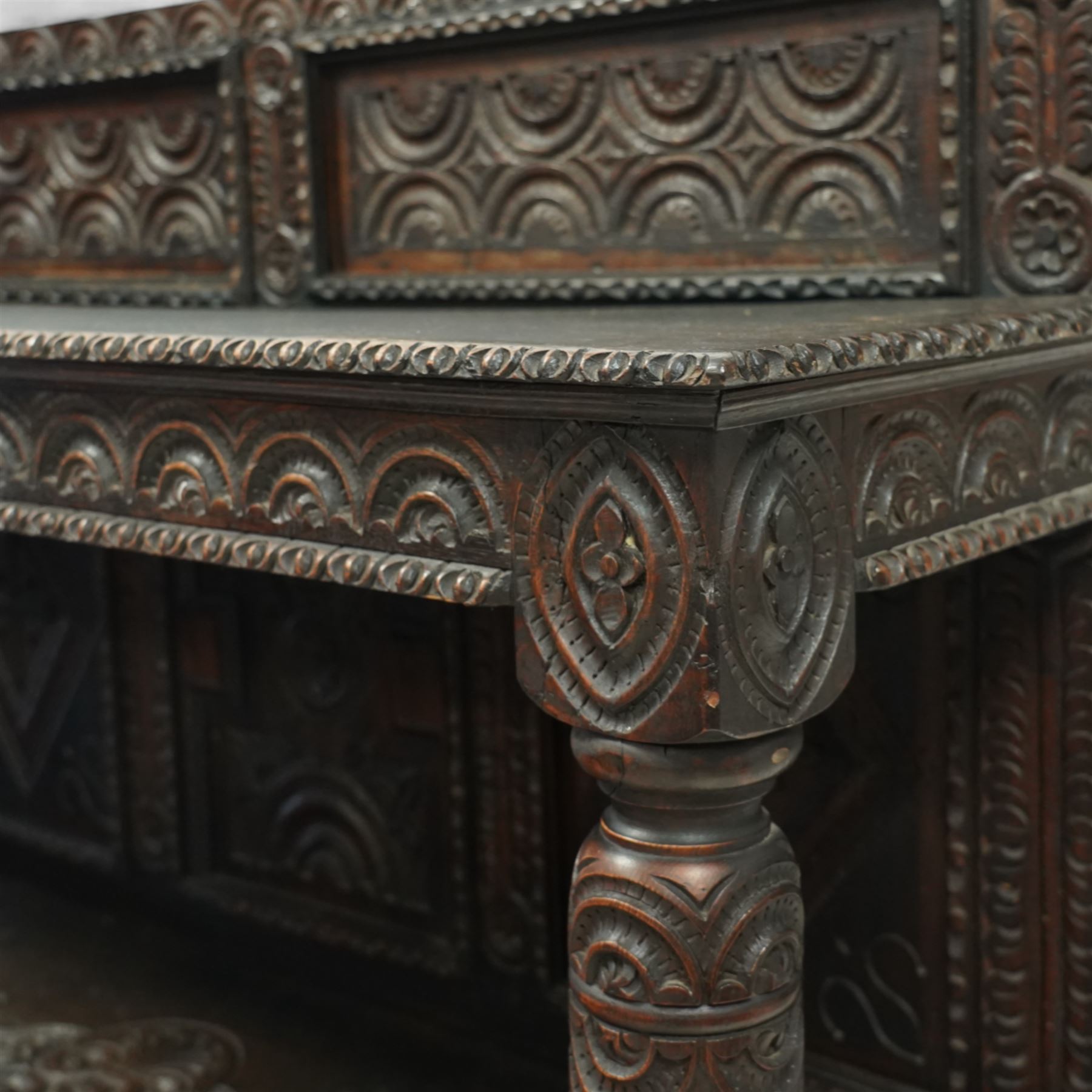 An early 20th-century Jacobean revival style carved oak Court Cupboard, tier-form of 17th-century design, rectangular form with carved back with geometric motifs and arches, projecting top section over twin baluster columns connecting the lower tier above a carved panelled front and panelled cupboard door with profusely carved lozenges and lunettes, above two base drawers, raised on block feet 