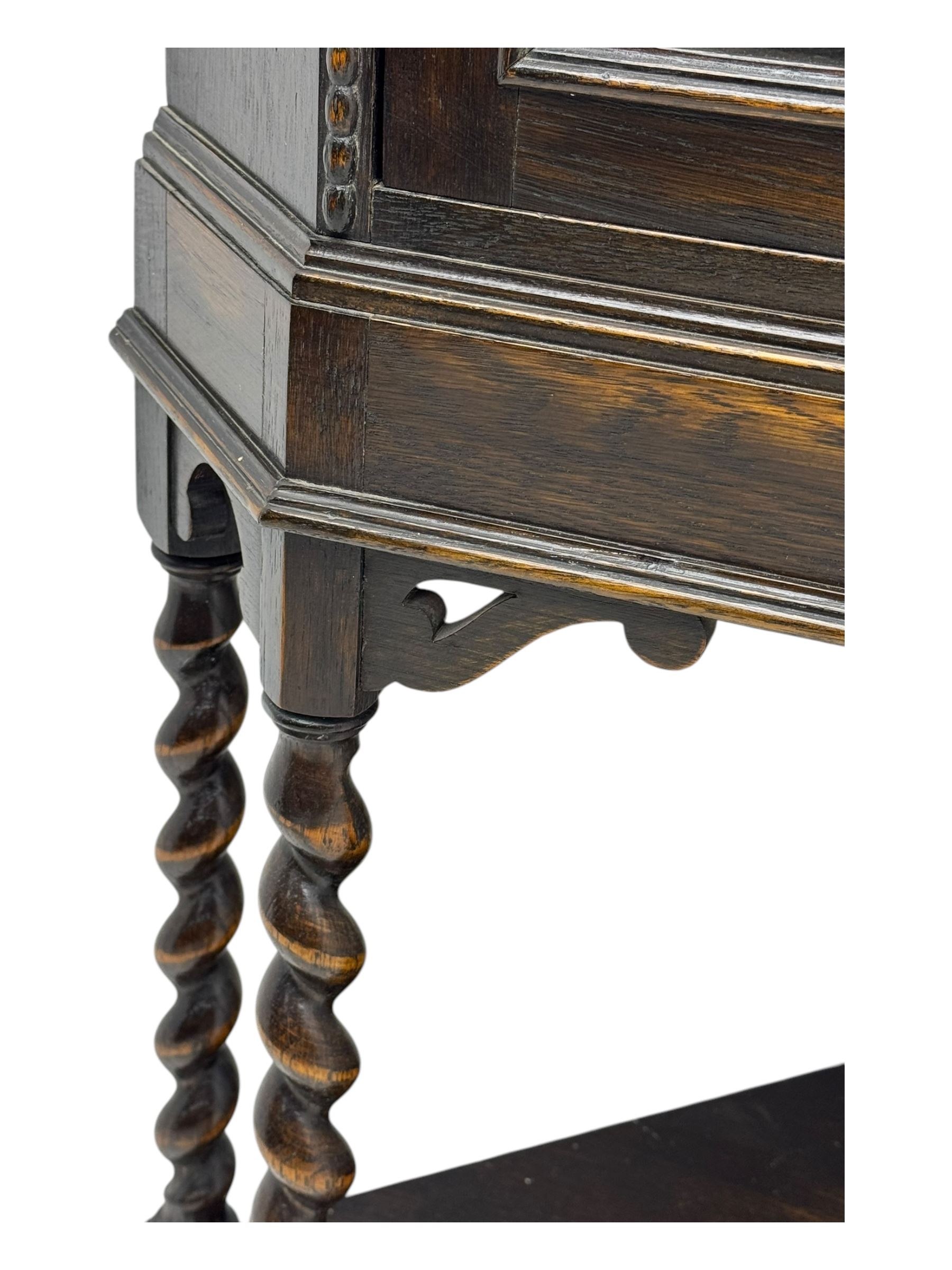 Early 20th century oak corner display cabinet, the moulded canted cornice over a single glazed door with geometric astragal bars, shelved interior, the lower tier forming a triangular platform, raised on four barley-twist supports united by shaped brackets