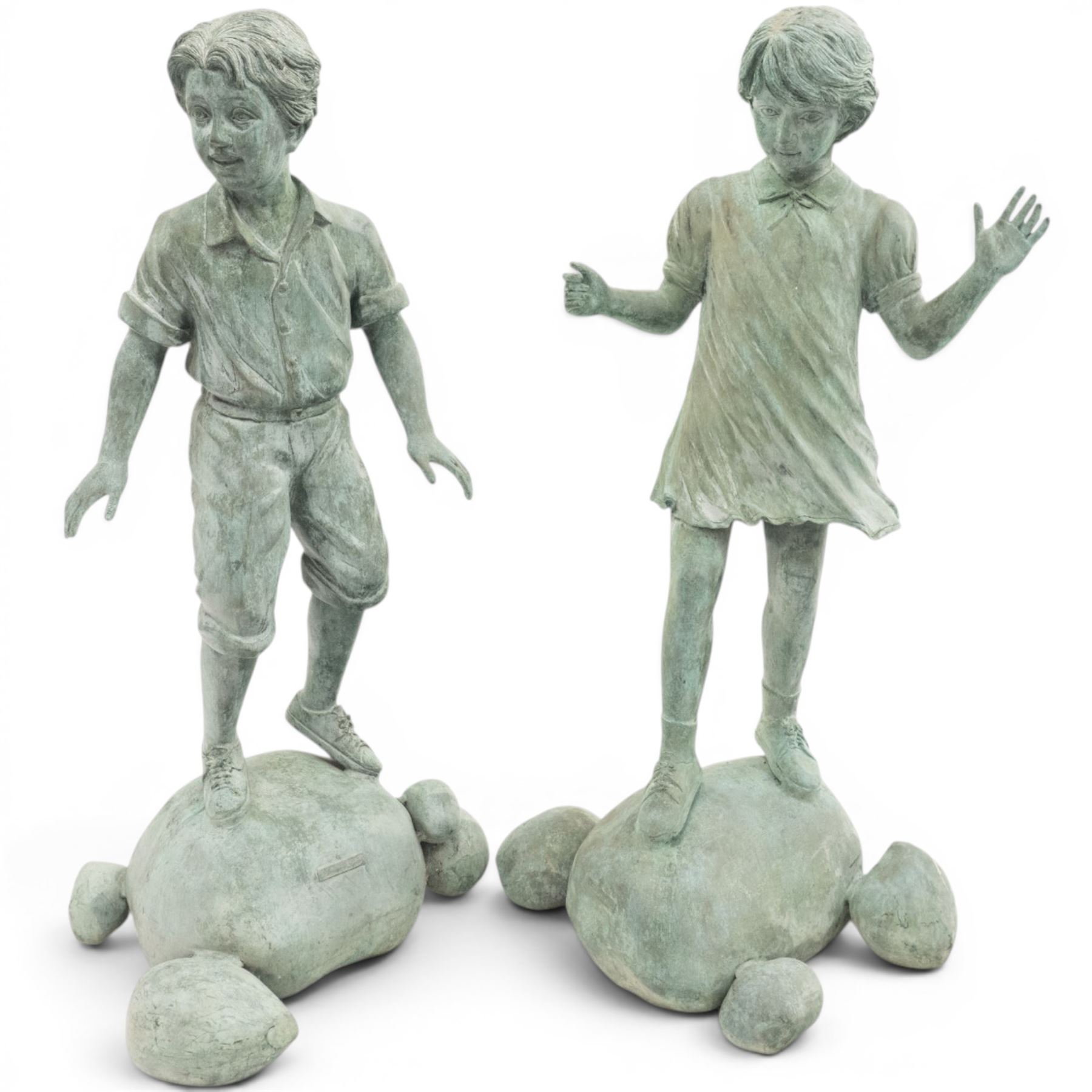 After Eneri Prosperi, bronze garden child statues, depicting children balancing on rock-form bases, the boy in shirt and shorts, the girl in dress with raised arms, surrounded by loose rounded pebble elements