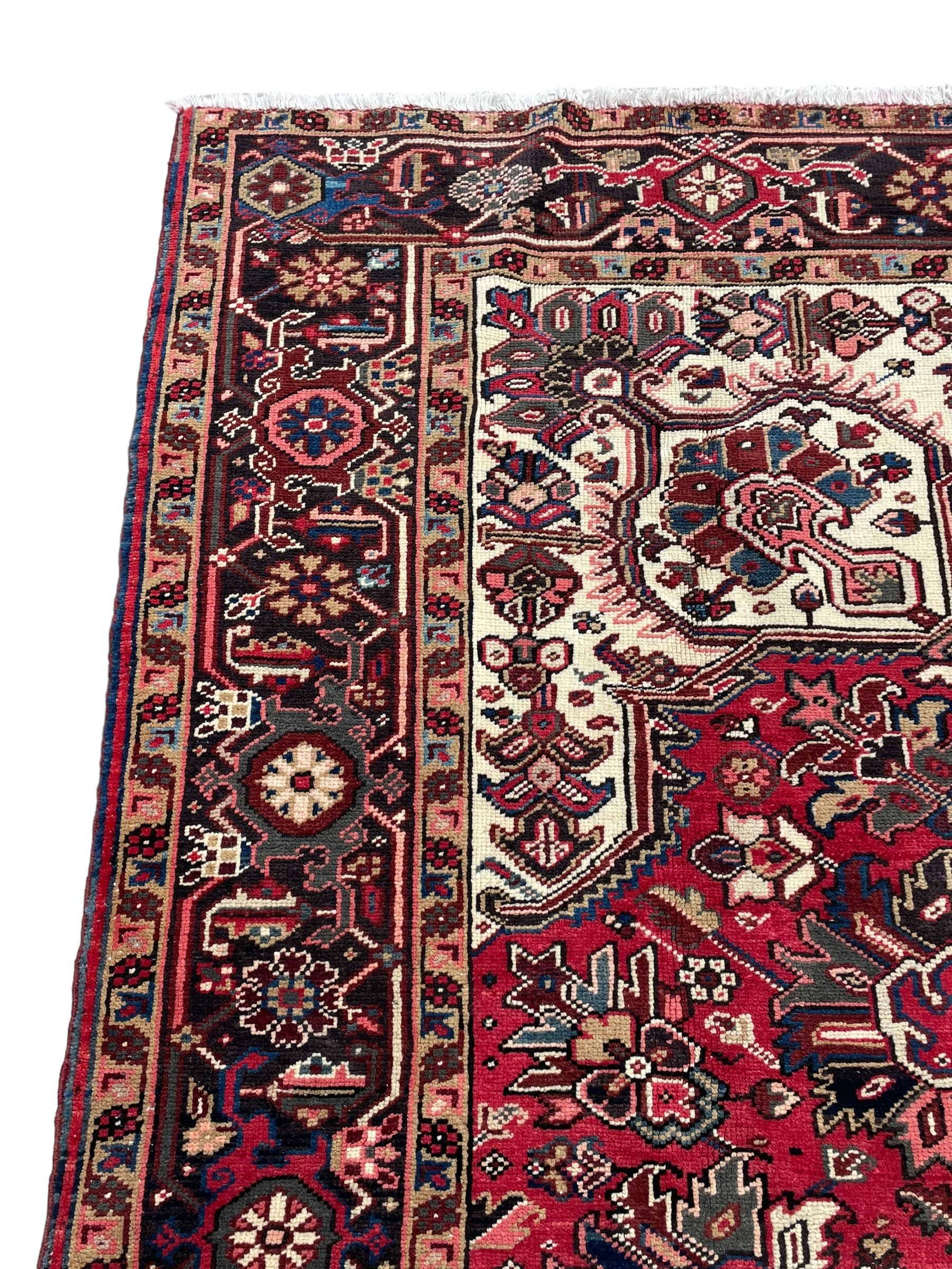 Persian Heriz red ground rug, central scrolled medallion on dark ground decorated with floral motifs, the surrounding field decorated with stylised plant motifs and smaller geometric symbols, guarded border with repeating pattern punctuated with rosettes and flower heads 
