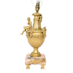 Pair of Rococo style gilt metal table lamps, the urn shaped bodies each with applied figure seated upon shoulder, and female mask to opposite side issuing scroll leading to top of neck, the body cast and applied with stiff leaf detail, and foliate swags and wreaths, upon square pink marble plinths with applied gilt metal mask mounts, and four gilt metal bracket feet, each supported a pleated cream silk closed top lampshade, H63cm