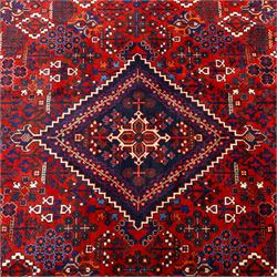 Central Persian Joshagen hand-knotted carpet, madder-red field with a central stepped lozenge medallion and densely filled with stylized geometric floral and tree motifs arranged in a repeating lattice enclosed by indigo spandrels and a broad navy border of angular palmette and vine ornament, flanked by multiple guard stripes, cotton fringe ends 