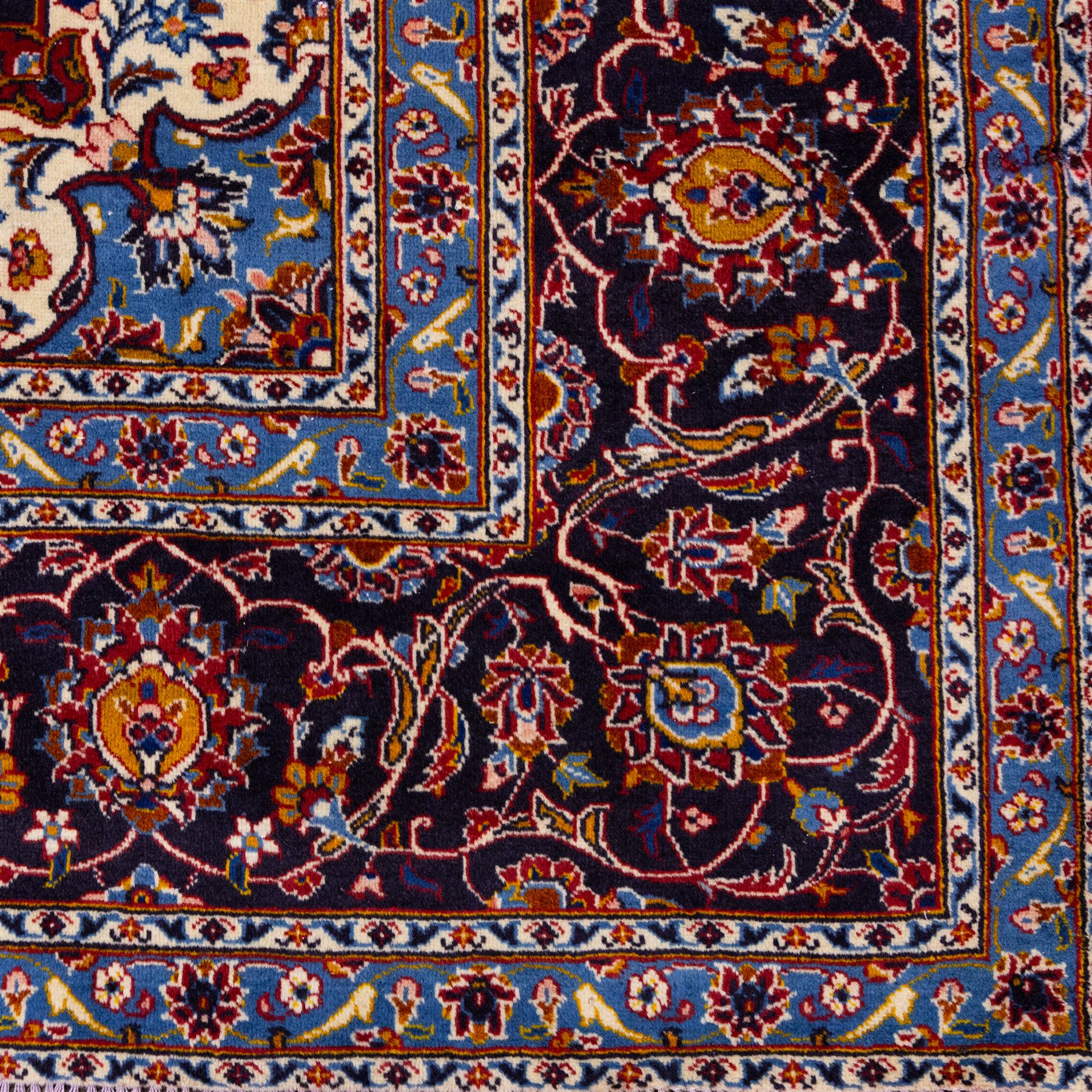 Central Persian Kashan hand-knotted carpet, rich crimson red field with a central cusped medallion with finely drawn floral sprays surrounded by dense scrolling vine work and palmettes, framed by a broad indigo ground border of flowering arabesques flanked by multiple guard stripes, in a palette of red, blue, ivory, sage green, and soft ochre tones, with cotton fringe ends    
