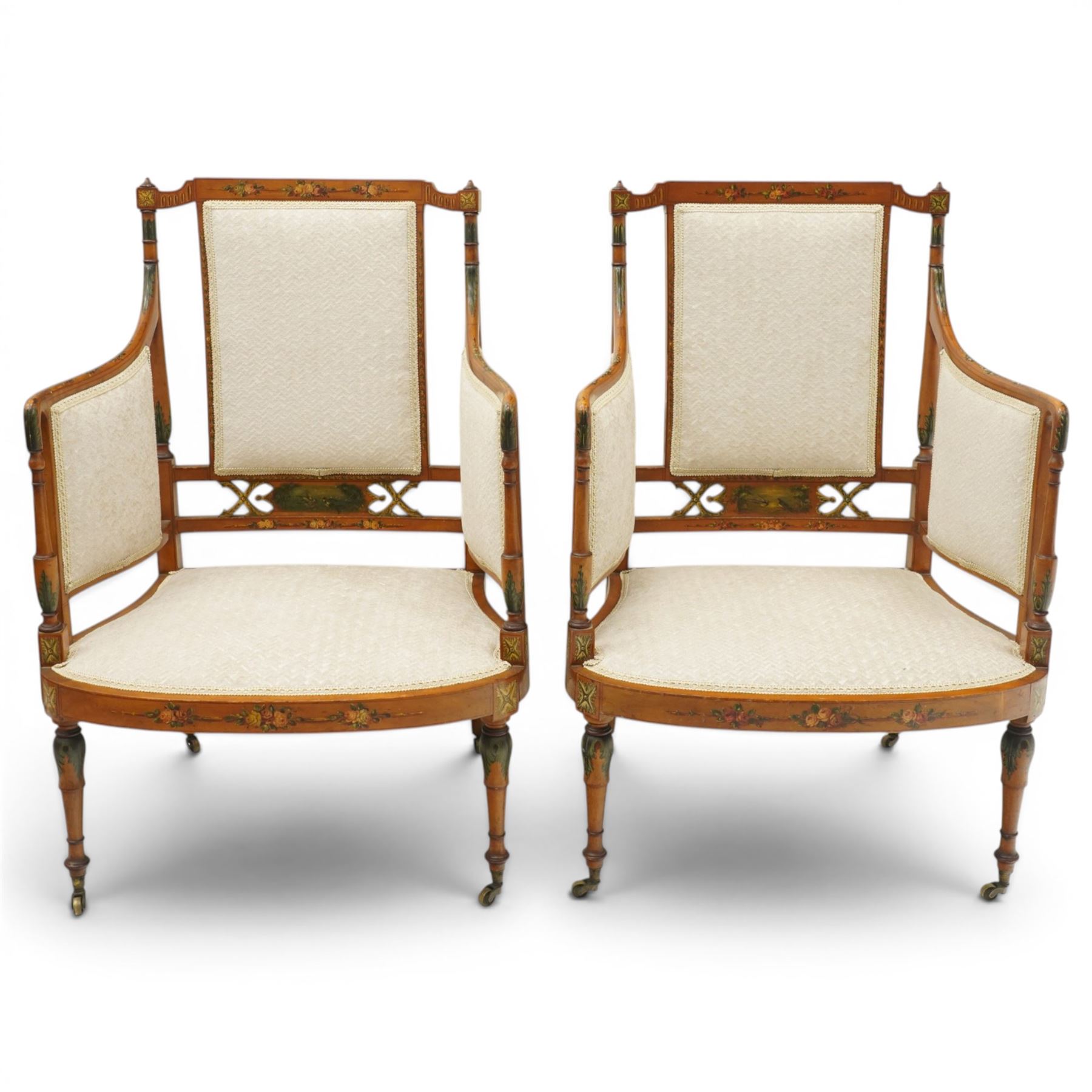 Pair of Victorian Sheraton Revival painted satinwood armchairs, the cresting rail painted with trailing floral bouquets flanked by illusion arcade motifs, over a cameo back and sides with acanthus painted swell-turned uprights, the lower back rail decorated with a bird scene and flanking pierced foliate design, upholstered in ivory fabric, raised on turned supports with further acanthus leaf paintwork, terminating to brass castors