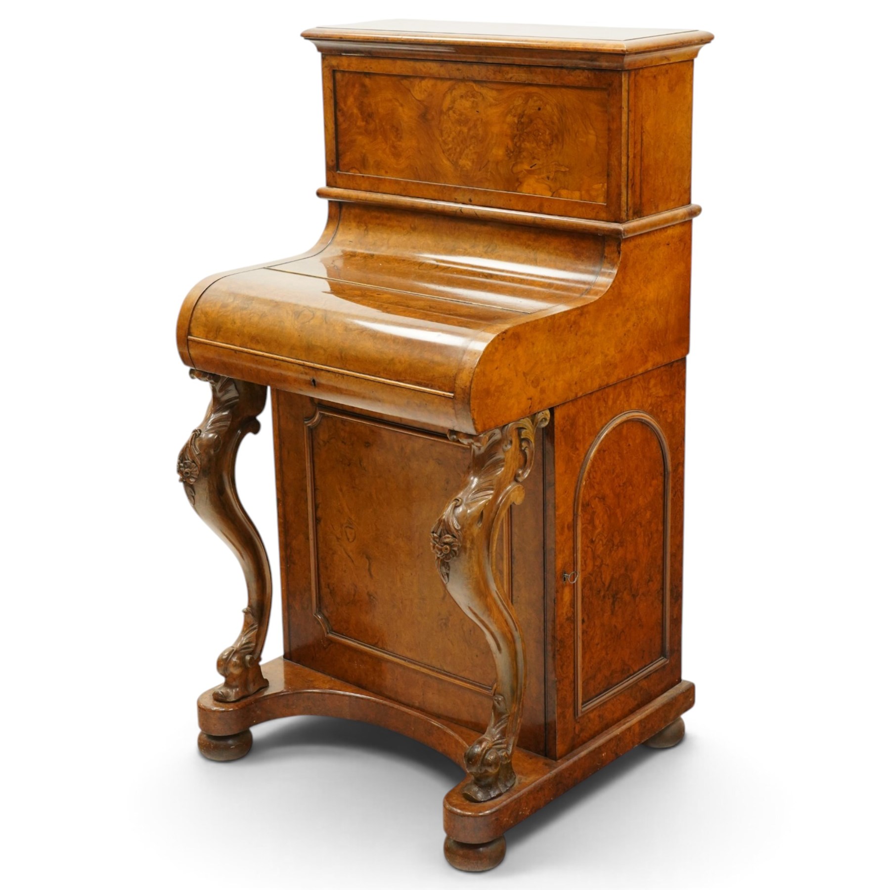 Victorian burr walnut piano-top Davenport, the raised rectangular superstructure with moulded cornice above a panelled fall-front enclosing drawers, the curved hinged writing slope opening to reveal a fitted interior above a sliding tooled leather writing surface, flanked by carved foliate scroll supports, the right side fitted with four drawers behind an arched panelled cupboard door with brass inset campaign-style handles, on a shaped plinth base and bun feet