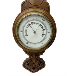 An English 1930’s solid oak carved hall barometer in a scroll shaped carved case with relief carving, aneroid movement, six-inch dial measuring barometric air pressure from twenty-six to thirty-one point nine inches, weather predictions in black upper and lower case gothic script with a blue steel indicating hand and brass recording hand, brass dial bezel with flat bevelled glass, mercury thermometer enclosed in a glazed rectangular box recording temperature in degrees Fahrenheit and Celsius on a white porcelain register.