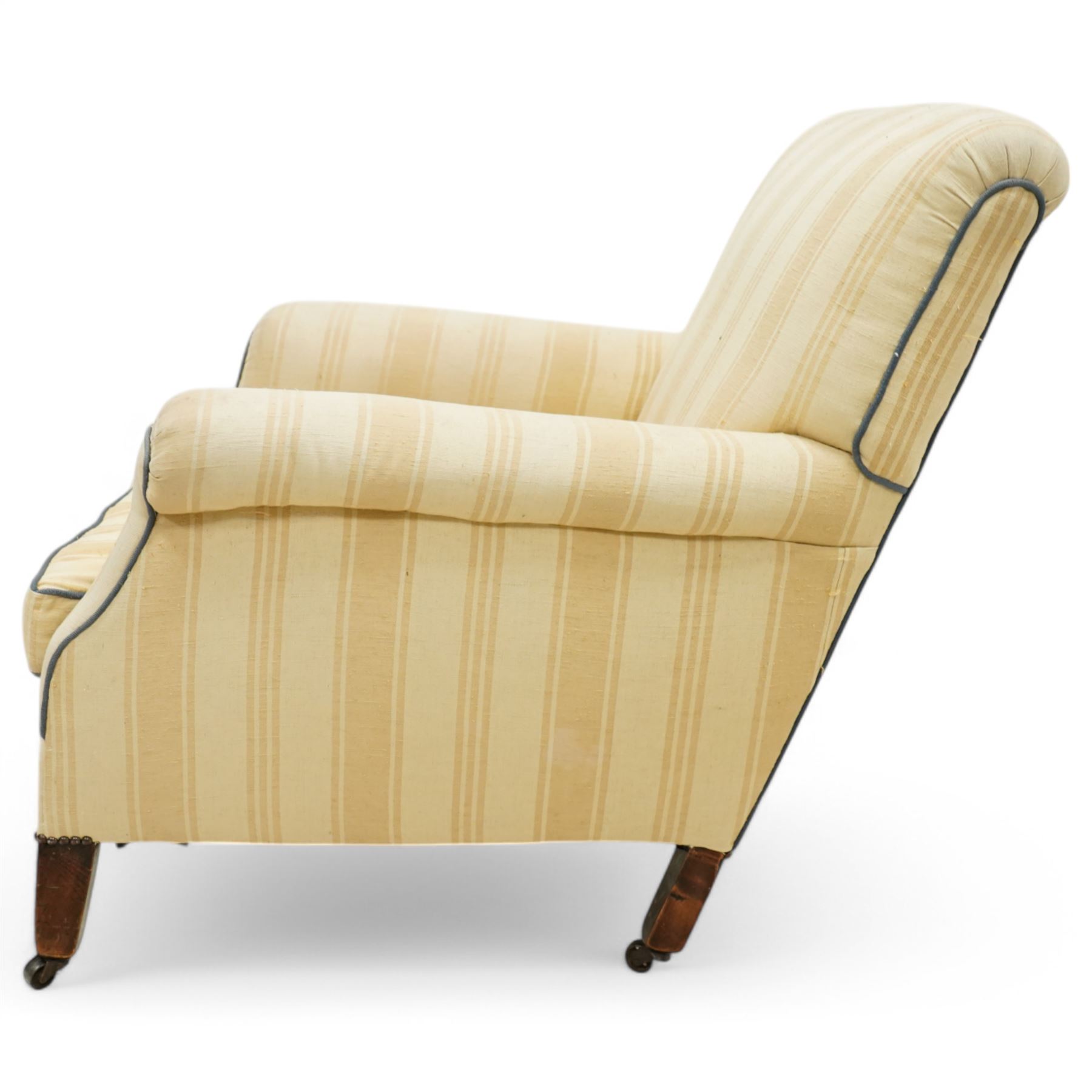 Early 20th century scroll arm armchair, upholstered in ivory striped cotton fabric with piped seams, sprung seat, on turned tapering front legs with brass caps and castors