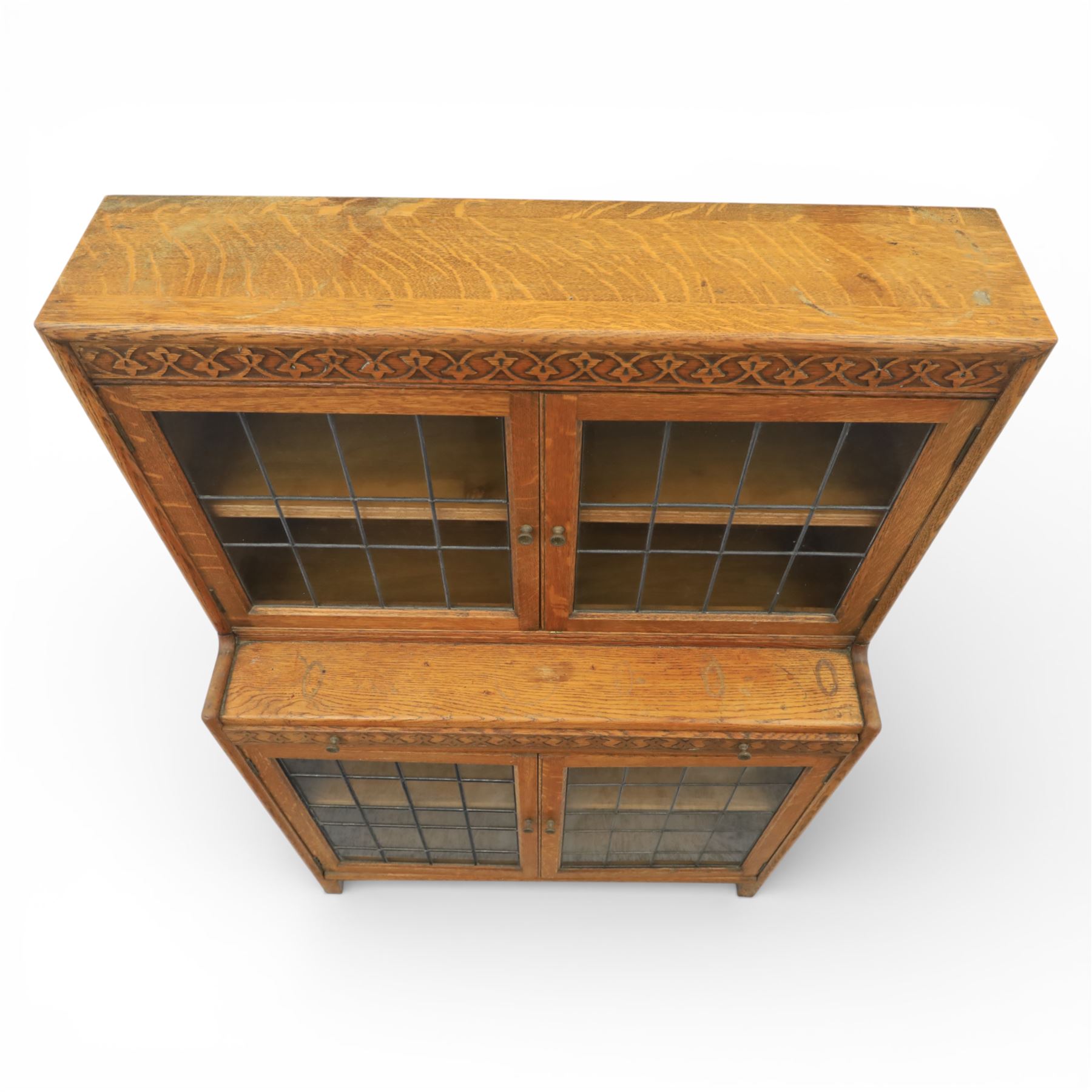 Early 20th-century Arts and Crafts glazed oak bookcase, upper section fitted with a pair of leaded glazed doors enclosing a fixed shelf above a projecting frieze drawer interior with baize-lined writing surface flanked by two compartments, carved with interlaced decoration, lower section fitted with a further pair of leaded glazed doors enclosing a single shelf, raised on short stile feet 