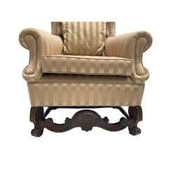 William and Mary style wingback armchair, upholstered in gold striped fabric, on walnut scroll carved supports joined by scroll and foliate carved middle rail, turned H-shaped stretchers, on castors