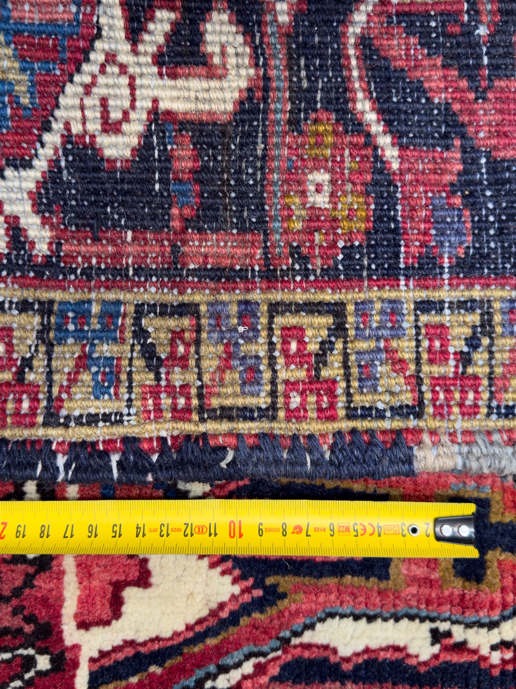 Northwestern Persian Heriz hand-knotted carpet, red madder field with a bold stepped medallion in indigo, ivory, and terracotta, flanked by angular floral motifs and stylized leaves, the spandrels set within a dark indigo border of rosettes and angular vine motifs between narrow guard stripes, 340cm x 245cm 