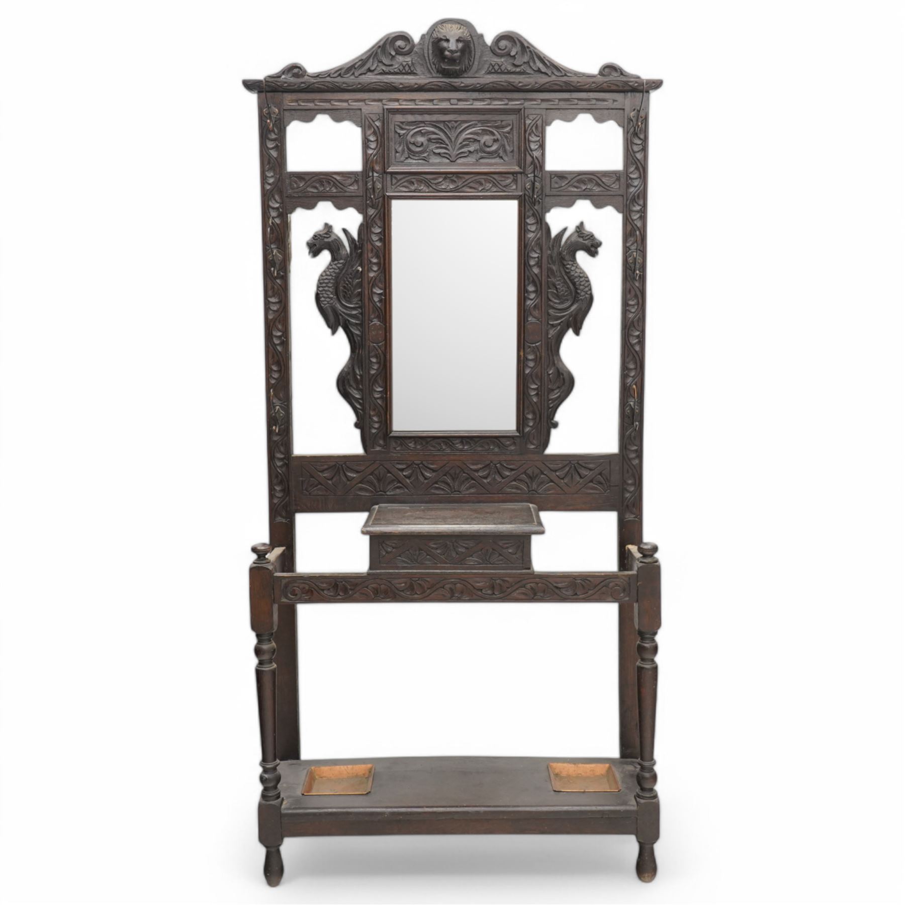 Late Victorian heavily carved oak hall stand, the scrollwork pediment with carved lion's head, bevelled mirror plate flanked by carved dragon motifs, over hinged storage compartment and umbrella stands with drip trays, on turned front supports