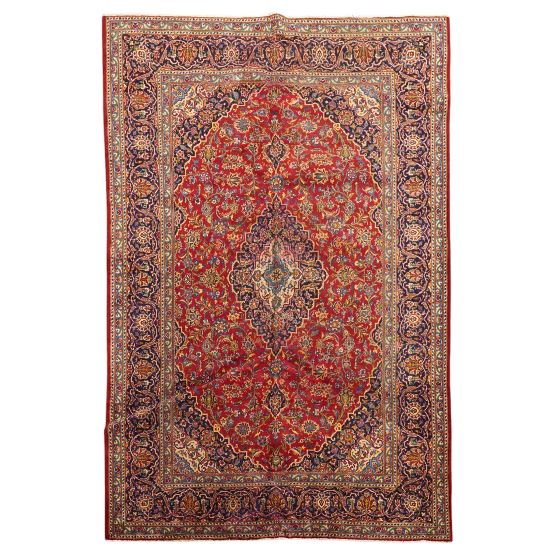 Persian Kashan hand-knotted carpet, madder red ground centred by shaped dark navy, ivory and pale blue floral medallion with pendants, surrounded by scrolling vines, palmettes and flowerheads, dark navy spandrels, enclosed by dark navy floral border with multiple floral guard stripes