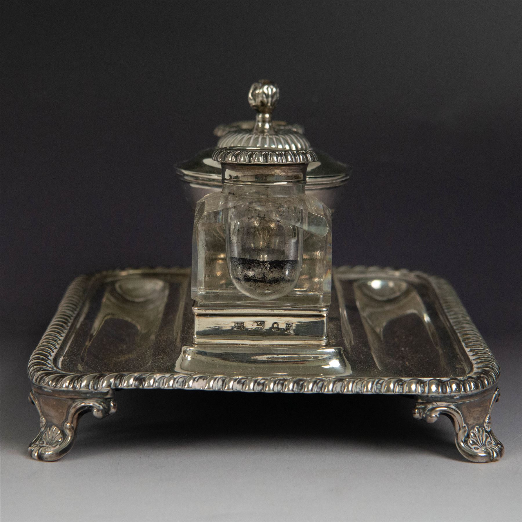 George IV silver desk stand, of rectangular form with rounded corners and oblique gadrooned rim, upon four embossed pad feet, with central pedestal dish of part fluted form with domed lid engraved with crest and with bud finial, with two recesses either side housing faceted glass inkwells, each with silver collars, hallmarked John & Thomas Settle, Sheffield 1825 & 1827, H11cm