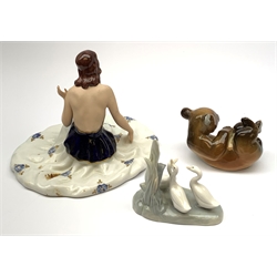 A Royal Dux figurine, modelled as a seated female figure with outspread gown, with pink triangle mark beneath, together with a Lomonosov figure of a recumbent bear, and a Nao figure group of three geese. (3). 