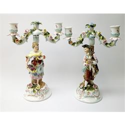 A pair of Continental two branch candlesticks, modelled with male figure and goat, and female fruit seller, the bases and candlestick branches encrusted with flowers, each H28cm.