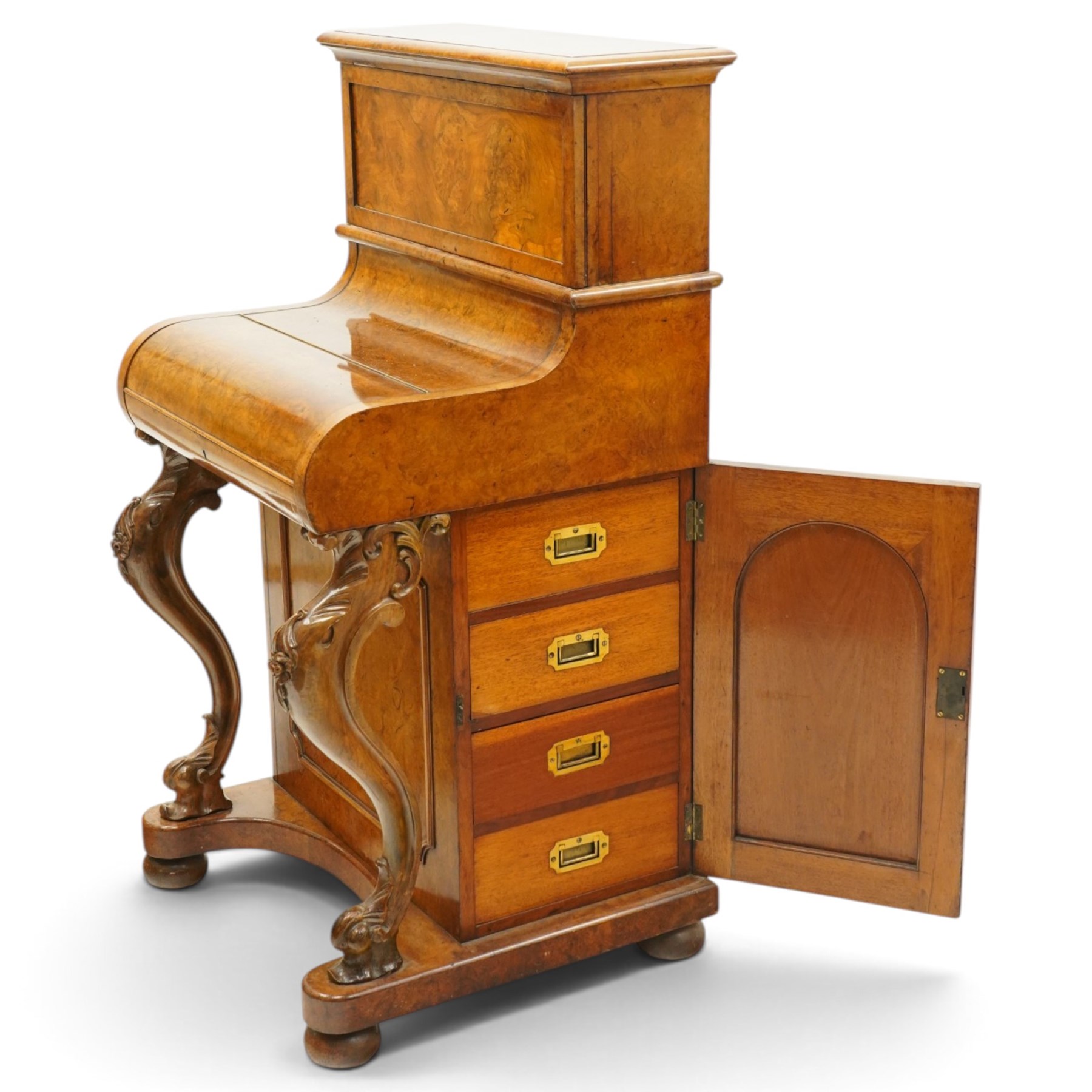 Victorian burr walnut piano-top Davenport, the raised rectangular superstructure with moulded cornice above a panelled fall-front enclosing drawers, the curved hinged writing slope opening to reveal a fitted interior above a sliding tooled leather writing surface, flanked by carved foliate scroll supports, the right side fitted with four drawers behind an arched panelled cupboard door with brass inset campaign-style handles, on a shaped plinth base and bun feet