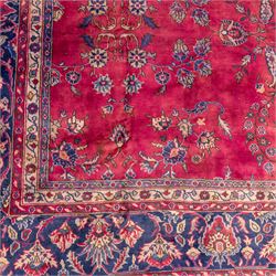 Large Persian-style mahal-type hand-knotted carpet, of rectangular form with a deep crimson field decorated with an all-over arrangement of stylized floral sprays and palmettes, with a central subtle medallion composition enclosed by a dark navy main border of scrolling floral motifs between narrow guard stripes with fringe ends 