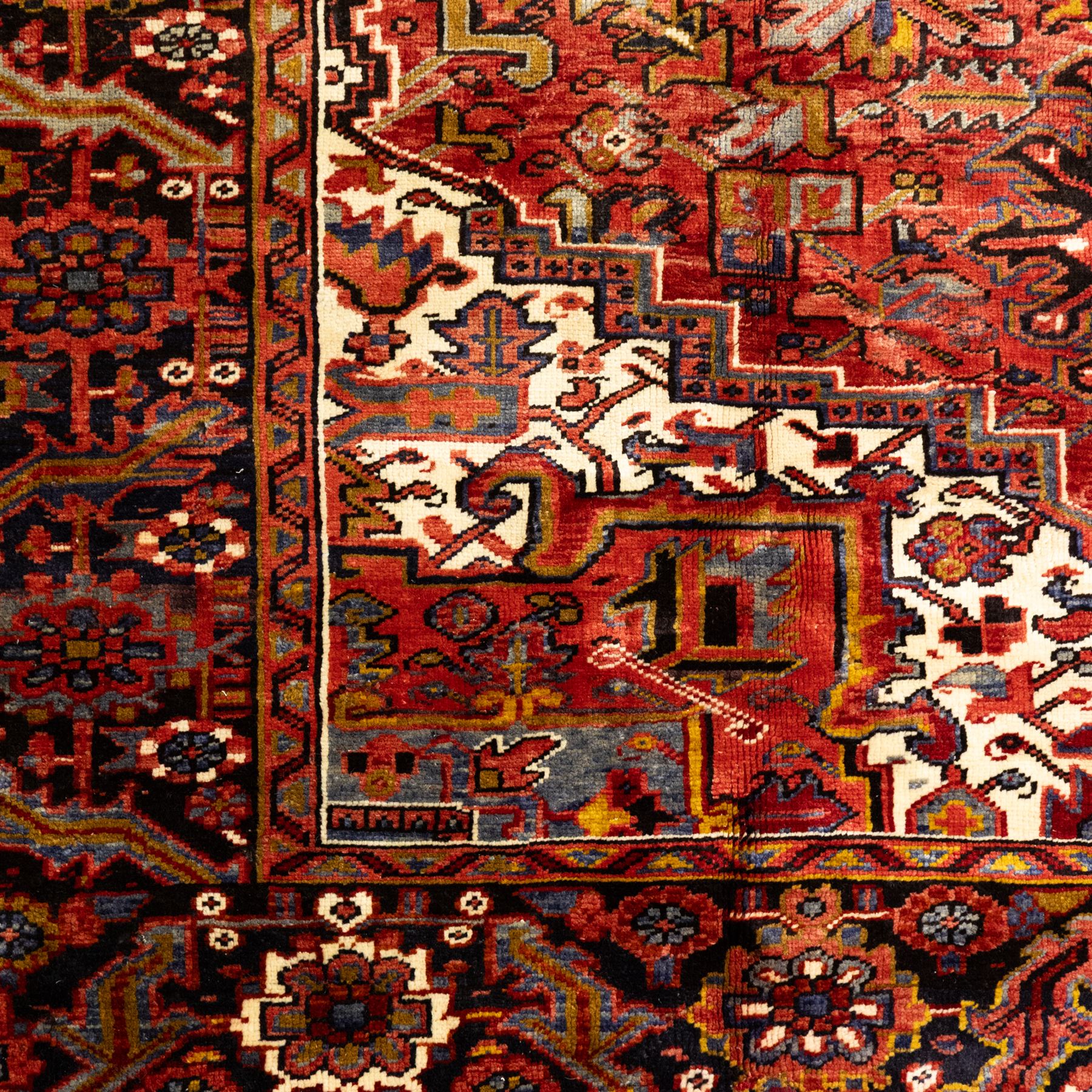 North West Persian Heriz hand-knotted carpet, madder red ground, with a large central geometric medallion with pendants, surrounded by angular foliate and floral motifs incorporating stylised rosettes, palmettes and serrated vine work, enclosed by ivory spandrels and a dark navy border with repeating geometric flowerheads and angular meandering vines, flanked by narrow patterned guard stripes, with cotton fringes