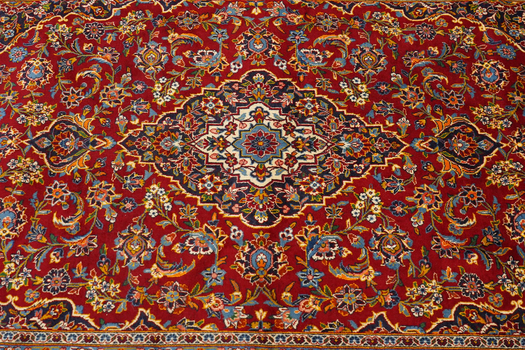 Central Persian Kashan hand-knotted carpet, crimson field with central cusped medallion in ivory, pale blue and indigo, issuing a network of scrolling arabesques, palmettes, and floral sprays, with conforming spandrels, main border of scrolling vinery and stylized palmettes, framed by multiple guard borders, with cotton fringe ends 