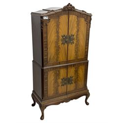 Early 20th century walnut veneered cocktail cabinet, shaped and carved foliate pediment above a pair of arched panelled doors with figured veneers and ornate cast escutcheons, interior is mirrored on the base and back, slide out mirrored shelf, flanked by foliate carved uprights, over a conforming lower cabinet, all raised on shaped apron and scroll carved cabriole feet