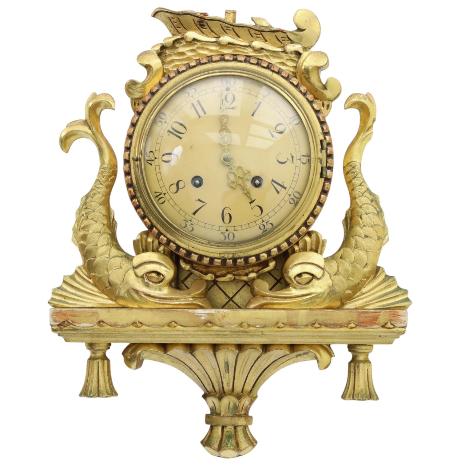20th century Continental giltwood wall clock, circular dial with Arabic numerals and pierced gilt hands, within a carved case surmounted by a scrolling crest and flanked by stylised dolphins, above a shaped shelf with tassel pendants and a foliate terminal, pendulum missing